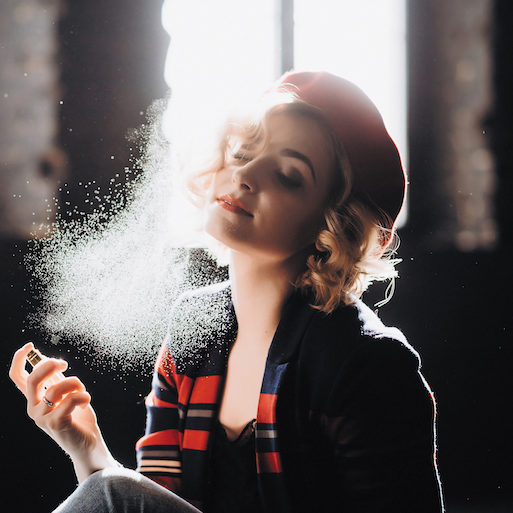 Een mooi blond meisje met een rode baret houdt een klein flesje in haar handen dat parfum op haarzelf spuit en geniet van haar armatuur. Tegenlicht, zachte focus.